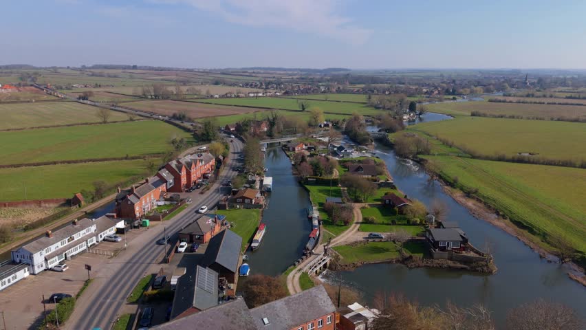 Aerial drone capture small village adjacent to large river with weir and river boats and busy road traffic in rural countryside of England on a sunny Spring day. Scenic picturesque life in England. 