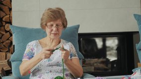 Elderly woman knitting with green yarn while sitting in a comfortable chair near a fireplace, focusing on her work. High quality 4k footage - Powered by Shutterstock - Get 15% off with code: PIKWIZARD15