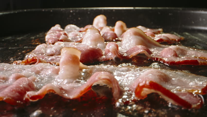 Frying pork bacon in pan side view close up slow motion. Fried slices of aromatic bacon are turned over in frying pan for better cooking. Start day with traditional breakfast. Aroma of bacon frying