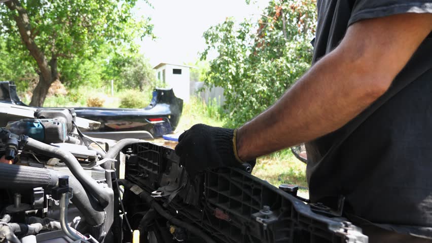 Male hands of repairman in gloves removing radiator from auto with tool outdoor. Arms of professional mechanic repairing vehicle at workplace. Adult male repairer making maintenance of car. Slow mo
