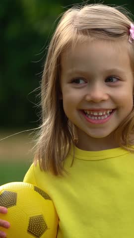 Portrait of little girl with yellow soccer ball looks at camera and laughs in summer park. Happy child waves hand outdoors. Football and sport. Vertical shot