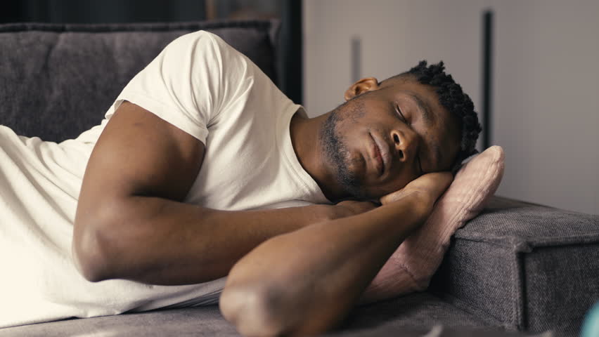 Tired African American male napping on the couch at home, taking a daytime rest