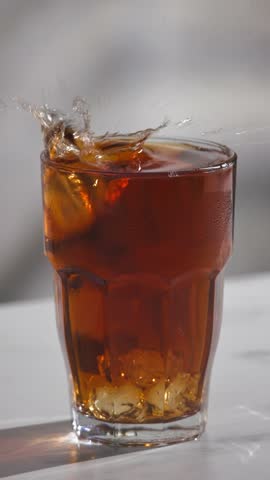 ice cube falls into a glass of iced tea, creating splashes on a gray kitchen table background in slow motion