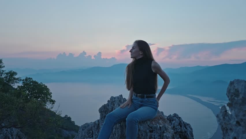 Stylish beautiful attractive young woman sits on a cliff, gazing at a sunset over mountains and sea, embodying a trend of mindful solitude and nature connection in a serene evening landscape.