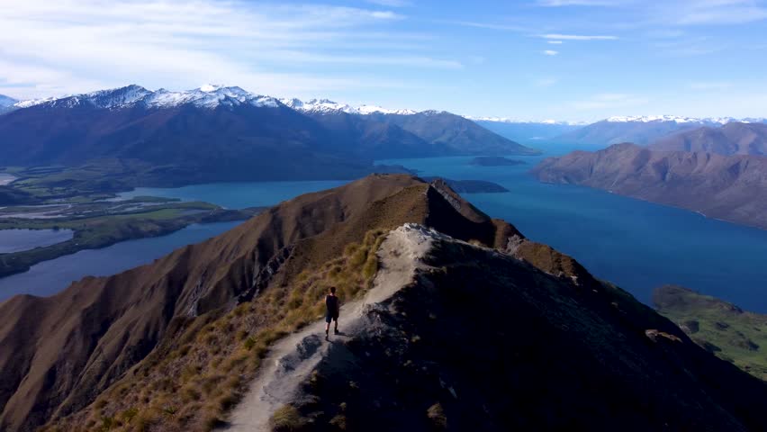 Drone view of young caucasian man walking towards a viewpoint and looking around with mountains and lakes in background on a sunny summer day at Roy's Peak, Wanaka, New Zealand.