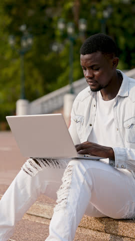 Portrait of African American man in denim outfit typing on laptop while sitting on stairs outdoors. Cheerful guy prepares online presentation on computer resting in park. Vertical video