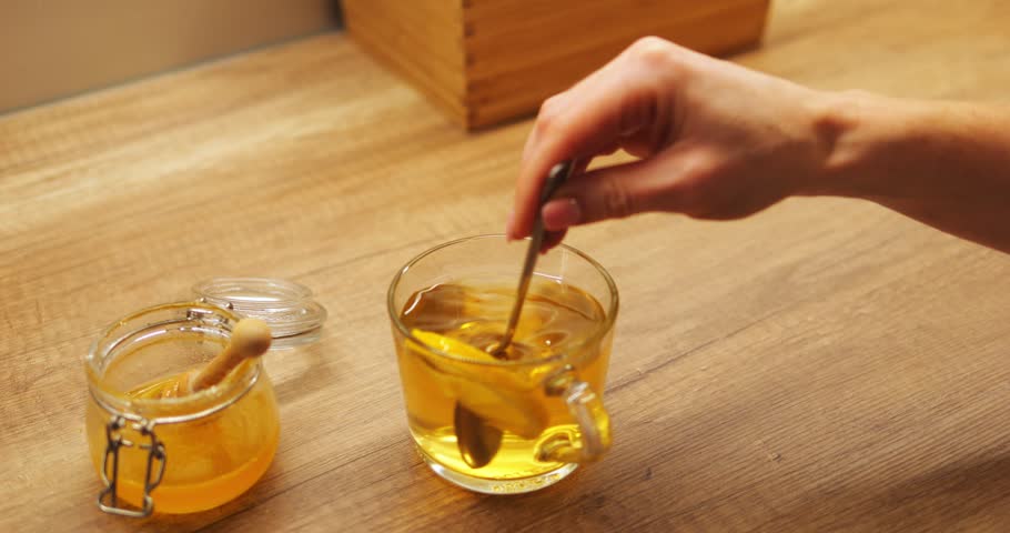 Woman hand stirs warm tea with lemon and honey in glass mug creating soothing beverage. Citrus slice twirls in boiling water creating refreshing flavor