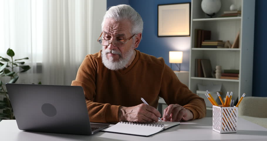 Senior man taking notes while learning online at desk indoors. Self-study