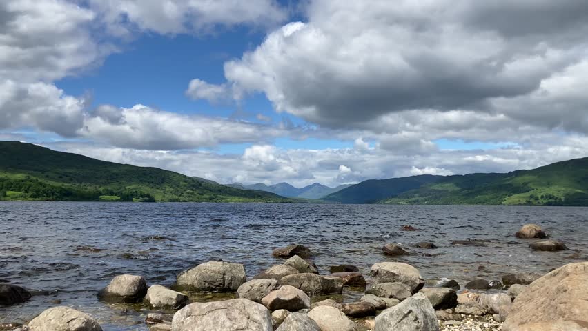 Scenic view of Loch Katrine in Scotland with rocks in the foreground, surrounded by lush green hills and mountains under a cloudy blue sky - Trossachs, UK