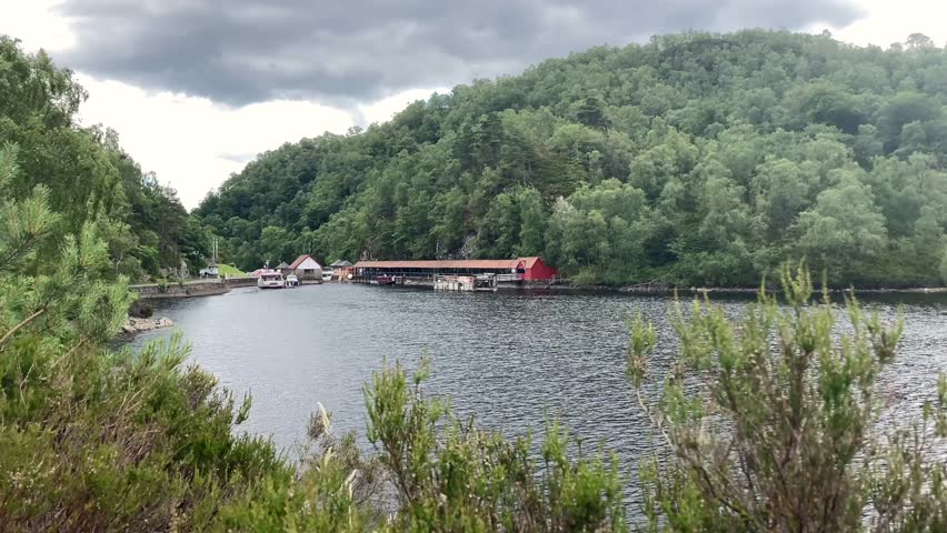 Scenic view of Loch Katrine in the Loch Lomond and Trossachs National Park, Scotland. A pier with boats and a forested hillside under a cloudy sky creates a tranquil atmosphere - UK