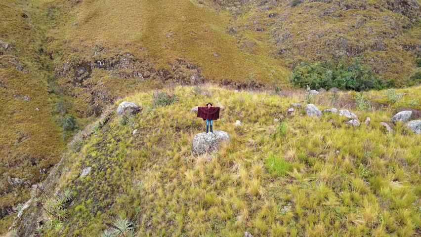 Young farmer wearing a poncho on a mountaintop, connecting with nature and rural tourism. Drone video of a young farmer wearing a hat under a panoramic sky. Quechua lifestyle.
