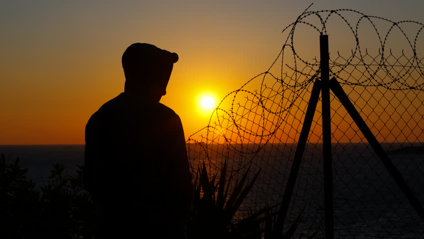 Man at sunset behind barbed wire. Hooded silhouette standing against sunset backdrop, removing hood while positioned near barbed wire fence, revealing personal identity against dramatic ocean horizon