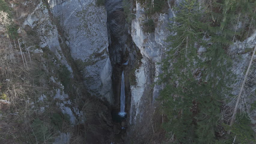 Tall waterfall cascading down a rocky cliff into a small pool. The steep canyon walls add a dramatic touch. Moss and vegetation grow on the rocks. A hidden natural gem, perfect for adventure seekers.