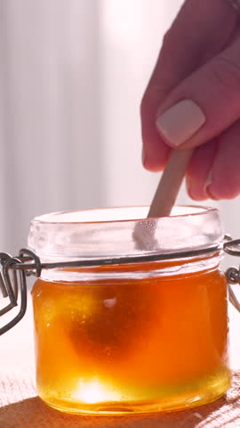Close-up of golden manuka honey slowly drizzling from a wooden dipper into a glass jar, captured in warm natural lighting with a soft curtain background for a cozy and organic kitchen scene.