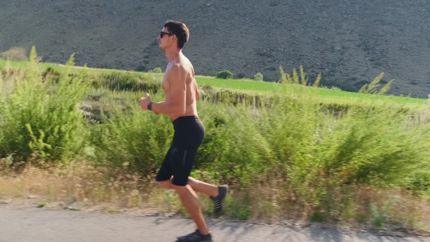 A muscular topless athlete in sunglasses runs a marathon along a flat road through hills and fields, his athletic build and rhythmic movements visible as he keeps pace, enjoying the warm sun.