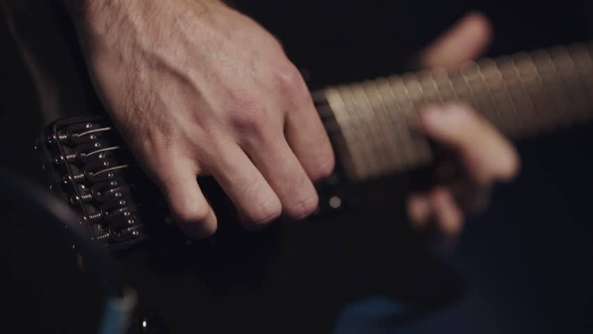 Close up slow motion capturing dynamic guitarist performing intense musical expression, revealing intricate hand movements across sleek black electric guitar against moody dark background