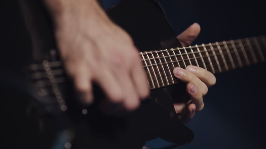 Close up slow motion capturing dynamic guitarist performing intense musical expression, revealing intricate hand movements across sleek black electric guitar against moody dark background