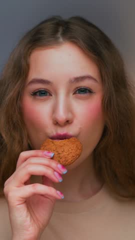 Portrait of Young Woman Eating Sweet Food and Looking at Camera. Real Person on Diet Consuming American Cookie as Lunch Snack. Playful Female Human Inside Cozy Living Room Alone with Sugary Fastfood