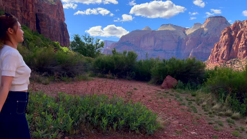 Asian young woman walking near majestic mountains enjoying picturesque scenery. Relaxing moment of travel, sunny day, clear blue sky, red rocks, lush greenery, peaceful adventure