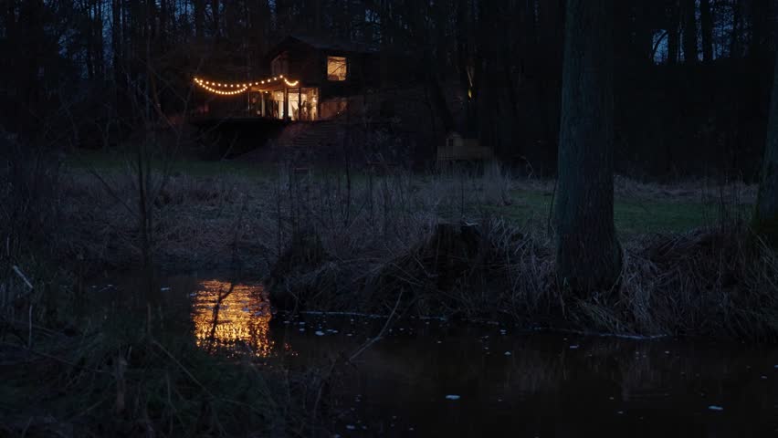 Illuminated wooden cottage in the middle of a forest by the river at autumn night