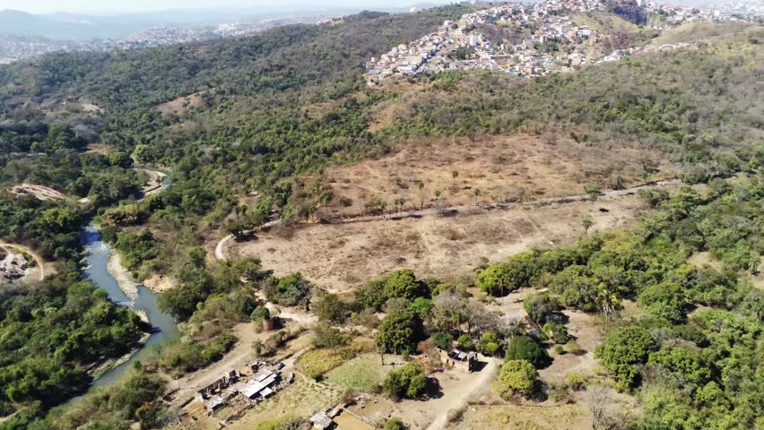 Aerial view of Izidora region in Belo Horizonte, Brazil, showcasing lush natural vegetation, farmland, winding roads, and a flowing creek amidst hills under clear skies. Nature meets urban fringe.