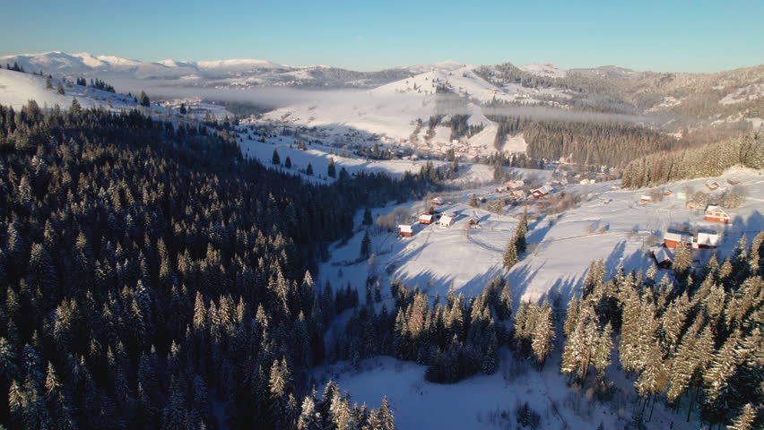 Aerial view of charming winter scene of snow-covered village nestled among rolling hills and dense forests. In distance, mist lingers over landscape, with mountains under clear blue sky.