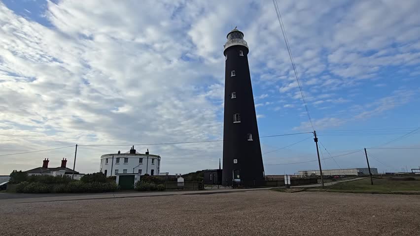 Wide-angle pan of the Old Lighthouse and adjacent Round House at Dungeness, Kent, UK, under a bright sky with power station in the background.