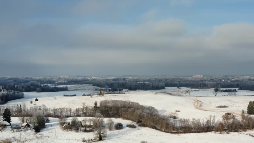 Drone shot of ancient windmill ruins on a snowy hilltop, surrounded by a frozen winter forest under the glowing sun. Aerial view over countryside landscape in winter.