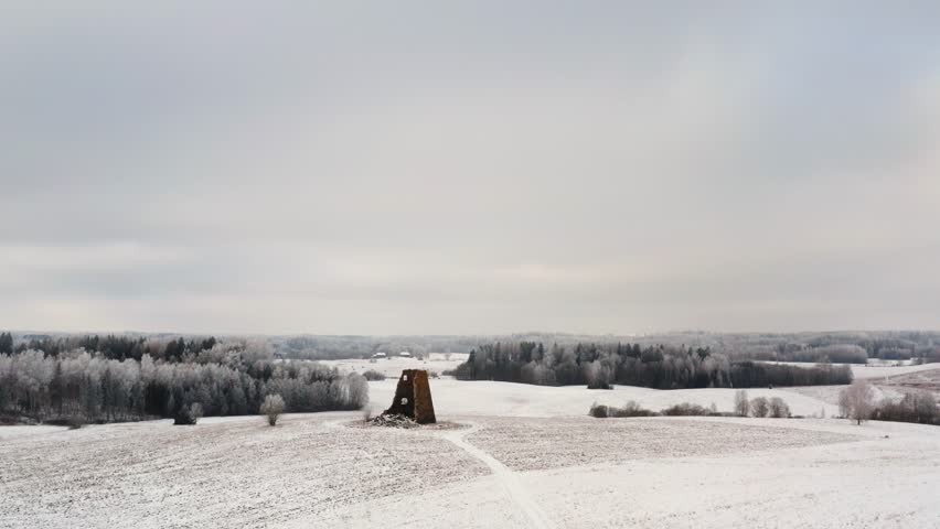 Aerial view of ancient windmill ruins on a snowy hilltop in the countryside with frozen forest in the background. Drone shot in a bright winter day.
