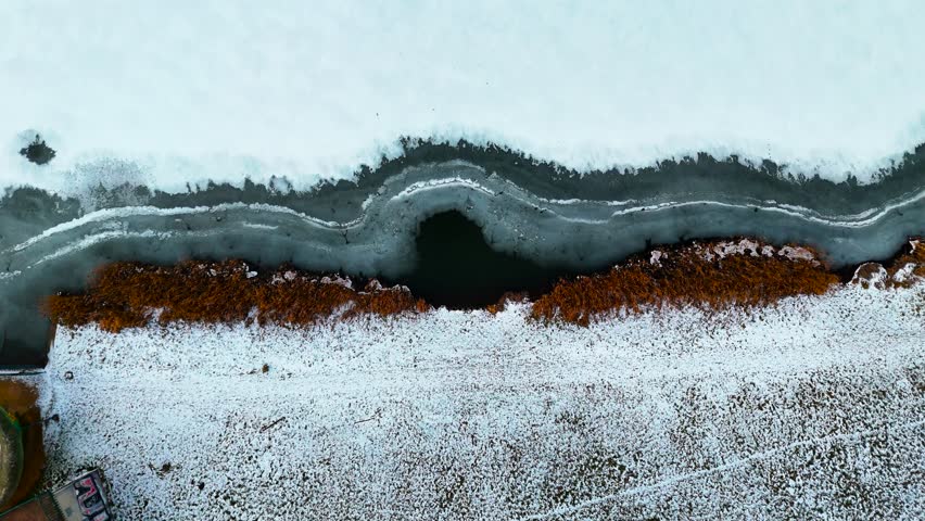 Aerial top-down descent over frozen lake shore with ice patterns and vegetation