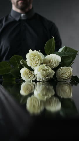 Vertical shot of priest finishing somber funeral ceremony while unrecognizable people placing roses on coffin, paying their respects