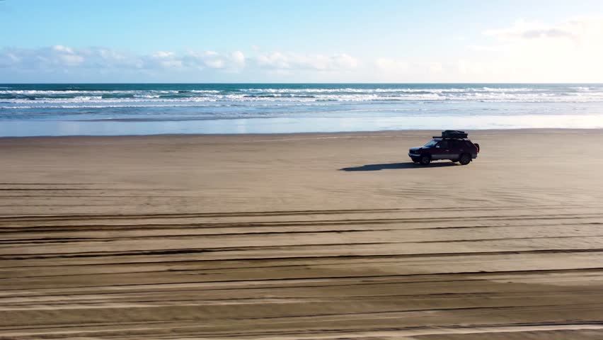 Drone view of offroad 4 wheel drive red car with rooftop tent driving on sand next to the ocean on a long beach on a sunny day at 90 Miles Beach, Northland, New Zealand.