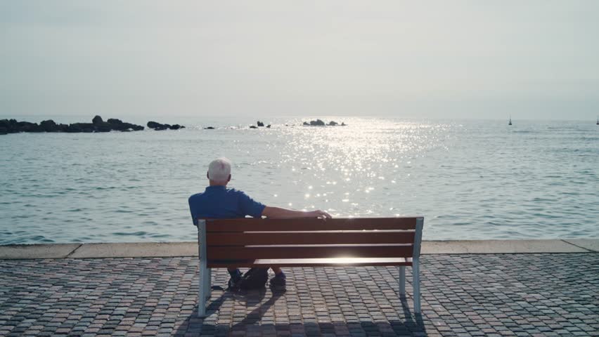 Old man sitting on a bench overlooking sea or ocean enjoying sunny day outside. Warm sun as senior male sits peacefully in a private quiet moment taking a break to look at view in vacation in Cyprus