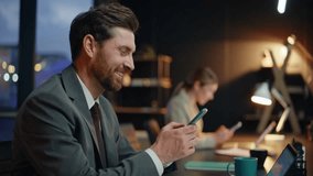 Evening coworkers messaging smartphones at lamp light workspace closeup. Two colleagues smiling each other sitting dark workplace with cellphones. Successful bearded businessman browsing mobile phone - Powered by Shutterstock - Get 15% off with code: PIKWIZARD15