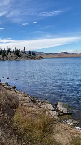 Waves of Clear Blue Water Splashing Against Rocky Shore With Mountain Forest Landscape Background and Bright Blue Sky