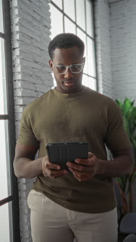 Handome man holding tablet in bright modern office interior with large windows and brick wall, wearing glasses and casual clothing, looking focused and professional.