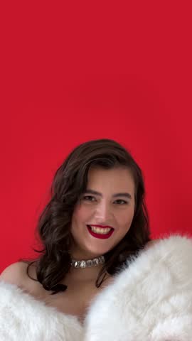 Vertical 4K video. Adult young white brunette pin up woman with bare shoulder, red lip, fluffy fur cape, diamond necklace smiles and looks at camera on scarlet. Copy space. Concept of 8 March