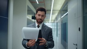 Bearded boss reviewing contracts walking contemporary office hall closeup. Smiling businessman in suit reading financial report on go. Positive manager flipping business documents waving hand indoors - Powered by Shutterstock - Get 15% off with code: PIKWIZARD15