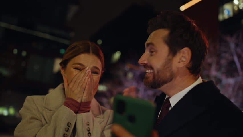 Overjoyed lovers standing night city reacting great news on smartphone closeup. Bearded businessman holding mobile phone sharing success with coworker lady. Two business owners reacting money win 
