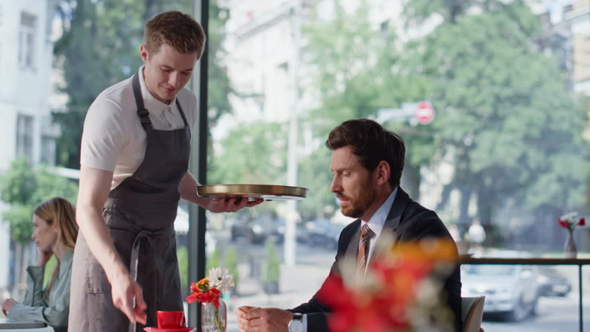 Restaurant visitor paying with credit card for coffee sitting table alone. Bearded businessman making cashless payment with pos terminal. Professional waiter taking empty cup from elegant man client.