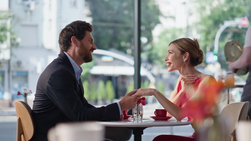 Lovely couple holding hands sitting cozy cafe. Smiling bearded man talking to beloved woman wearing red dress spending date in restaurant. Boyfriend confessing in love to beautiful girlfriend at table
