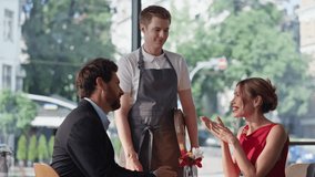 Friendly waiter taking order in happy romantic couple at modern cafeteria. Smiling bearded man talking to beautiful woman wearing red dress. Pair in love dating in cozy restaurant ordering drinks. - Powered by Shutterstock - Get 15% off with code: PIKWIZARD15
