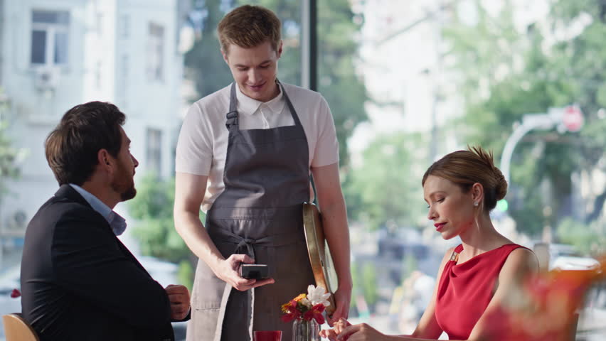Romantic pair sitting restaurant enjoying time together. Resting couple paying cafe bill separately by cashless transaction. Friendly waiter bringing pos terminal to spouses for contactless payment.