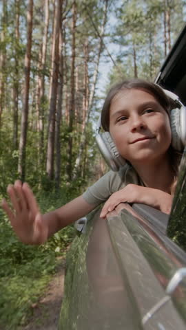 Vertical shot of happy young boy in headphones leaning out of car window, moving his hand and enjoying view of scenic woods during road trip