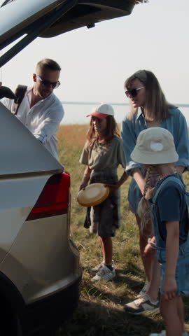 Vertical shot of family walking to car, loading belongings into trunk while leaving sunny lakeshore after camping trip