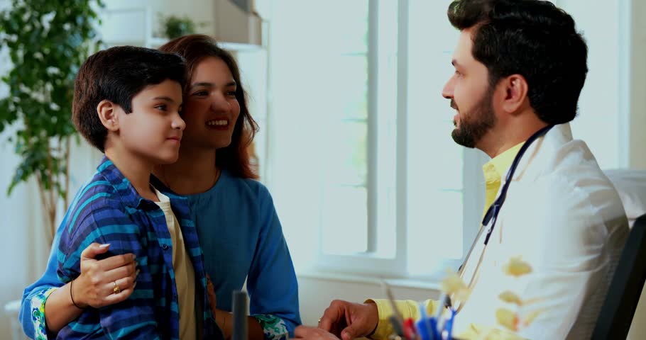 Indian Asian male doctor using stethoscope to check young boy’s general symptoms in modern clinic, concerned mother watches examination in professional medical setting at hospital, healthcare facility