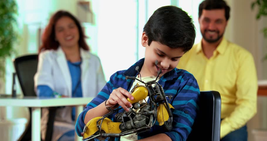Indian Asian playful small boy sitting on chair, happily holding a motorcycle toy while looking at camera in a friendly medical clinic, hospital, with caring lady doctor, father in the background