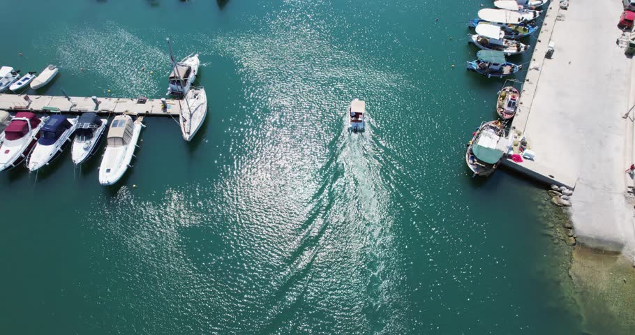 Small fishing boat sail among boats and yachts docked alongside a pier, with clear blue waters sparkling in the sunlight