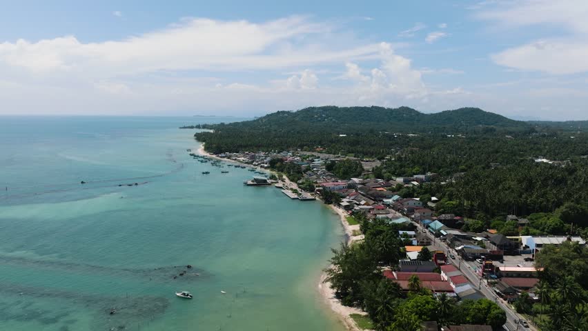 Coastal village surrounded by dense tropical vegetation and clear turquoise sea under a bright sky. Ko Samui, Thailand.