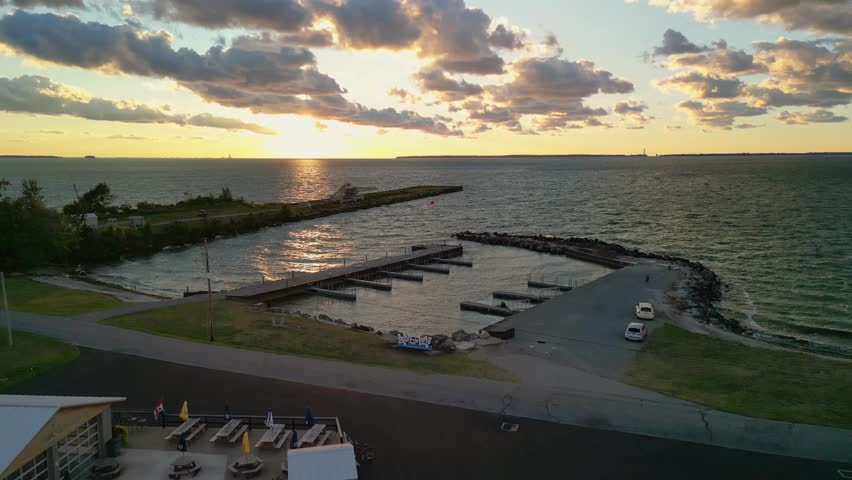 Aerial drone shot of a marina at sunset, with golden sunlight reflecting off the calm waters, a long pier extending into the lake, and a dramatic sky filled with clouds